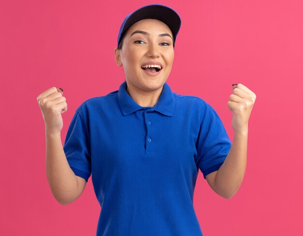 Repartidor joven en uniforme azul y gorra mirando al frente apretando los puños feliz y emocionado de pie sobre la pared rosa