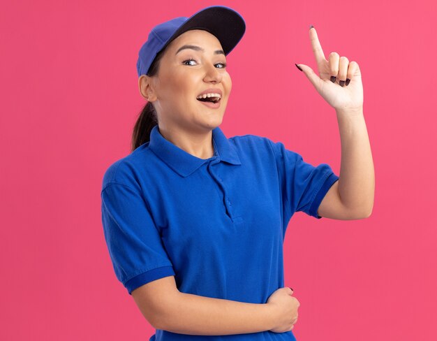 Repartidor joven en uniforme azul y gorra mirando al frente feliz y positivo mostrando el dedo índice con nueva idea de pie sobre la pared rosa