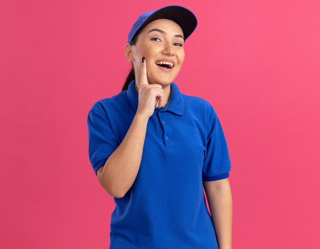Repartidor joven en uniforme azul y gorra mirando al frente sonriendo alegremente feliz y positivo de pie sobre la pared rosa