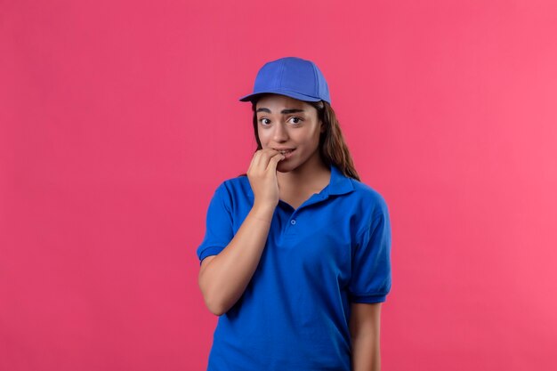 Repartidor joven en uniforme azul y gorra mirando a la cámara nervioso y estresado morderse las uñas de pie sobre fondo rosa