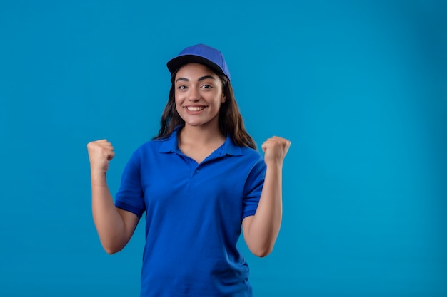 Repartidor joven en uniforme azul y gorra mirando a la cámara sonriendo alegremente levantando el puño regocijándose de su éxito y victoria de pie sobre fondo azul