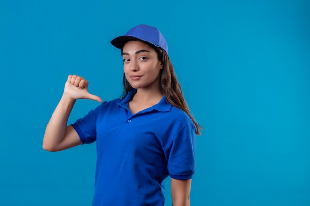 Repartidor joven en uniforme azul y gorra mirando confiada apuntando a sí misma con el pulgar de pie satisfecho y orgulloso sobre fondo azul