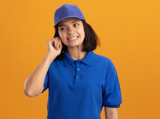 Repartidor joven en uniforme azul y gorra mirando a un lado confundido parado sobre pared naranja