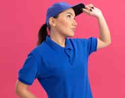 Foto gratuita repartidor joven en uniforme azul y gorra mirando a un lado desconcertado parado sobre pared rosa