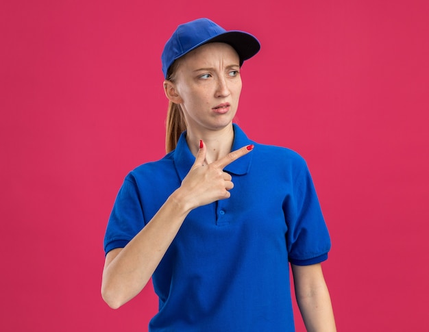 Repartidor joven en uniforme azul y gorra mirando a un lado con expresión escéptica apuntando con el dedo índice hacia el lado