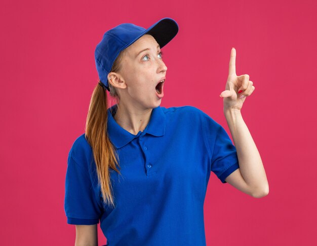 Repartidor joven en uniforme azul y gorra mirando sorprendido y sorprendido apuntando con el dedo índice a algo parado sobre la pared rosa