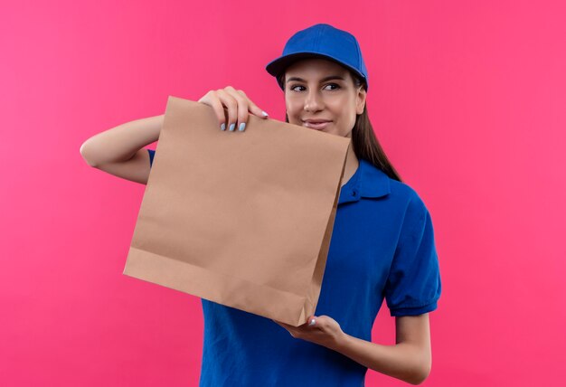 Repartidor joven en uniforme azul y gorra mostrando paquete de papel mirando a un lado sonriendo amable