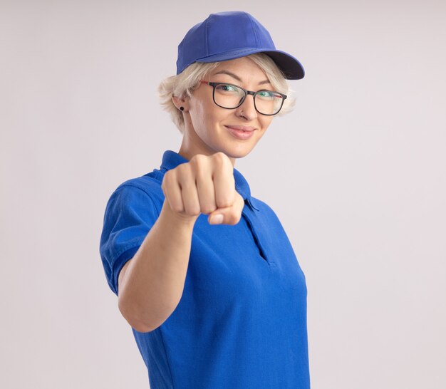 Repartidor joven en uniforme azul y gorra mostrando puño sonriendo confiado de pie sobre la pared blanca