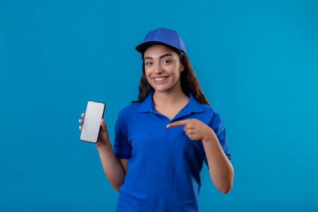 Repartidor joven en uniforme azul y gorra mostrando smartphone apuntando con el dedo a él sonriendo amable de pie sobre fondo azul.