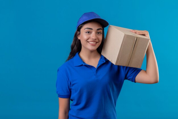 Repartidor joven en uniforme azul y gorra con paquete de caja dárselo a un cliente de pie sobre fondo azul.