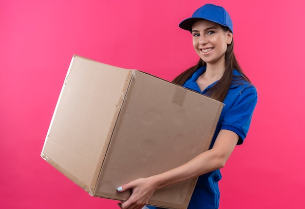 Repartidor joven en uniforme azul y gorra con paquete de caja grande mirando a la cámara con una sonrisa en la cara