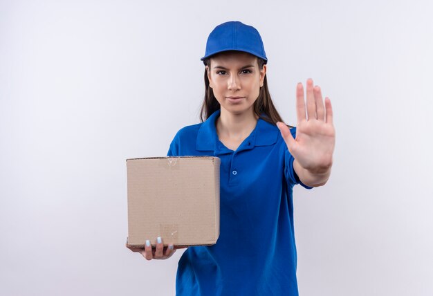 Repartidor joven en uniforme azul y gorra con paquete de caja haciendo señal de stop con la mano mirando a la cámara con cara seria