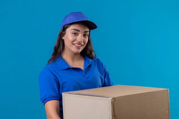 Repartidor joven en uniforme azul y gorra con paquete de caja mirando a la cámara sonriendo confiada feliz y positiva de pie sobre fondo azul.