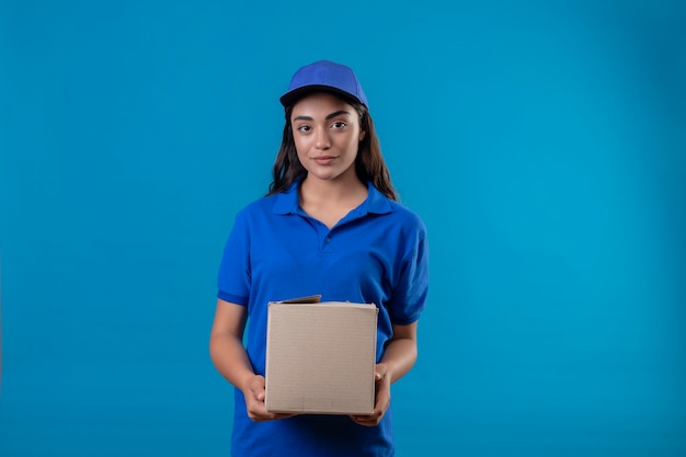 Repartidor joven en uniforme azul y gorra con paquete de caja mirando a la cámara sonriendo confiada feliz y positiva de pie sobre fondo azul.