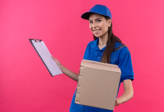 Foto gratuita repartidor joven en uniforme azul y gorra con paquete de caja y portapapeles mirando confiado sonriendo