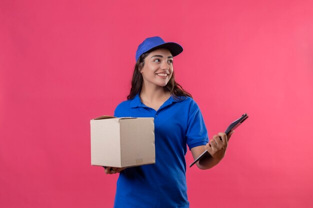 Repartidor joven en uniforme azul y gorra con paquete de caja y portapapeles mirando a un lado sonriendo alegremente de pie sobre fondo rosa