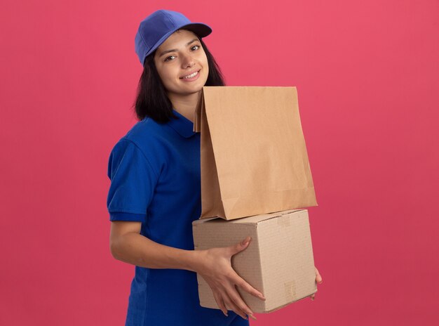 Repartidor joven en uniforme azul y gorra con paquete de papel y caja de cartón sonriendo con cara feliz de pie sobre la pared rosa