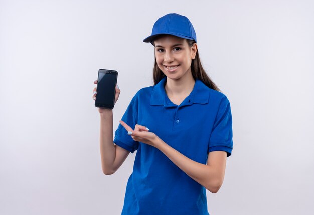 Repartidor joven en uniforme azul y gorra presenta smartphone mirando a cámara con sonrisa en la cara