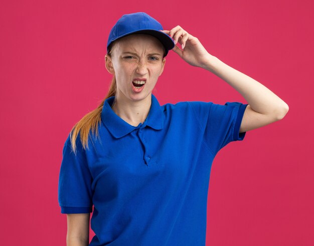 Repartidor joven en uniforme azul y gorra que se confunde con la mano en la cabeza por error de pie sobre la pared rosa