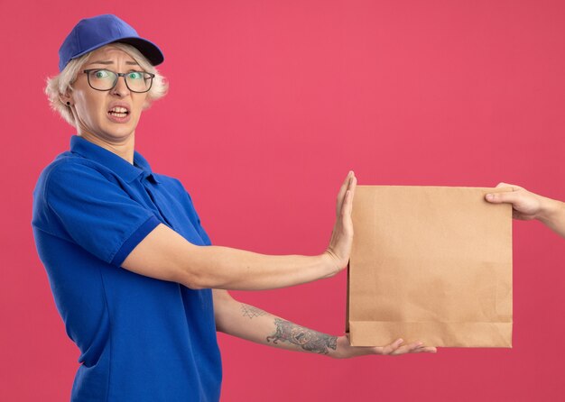 Repartidor joven en uniforme azul y gorra que se niega a tomar un paquete de papel con aspecto confundido y preocupado de pie sobre la pared rosa