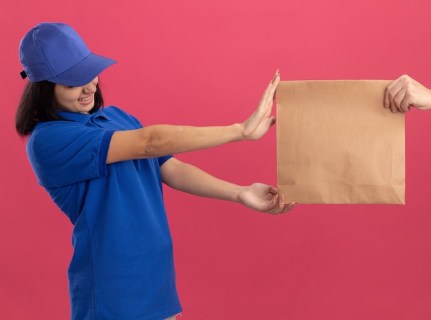 Repartidor joven en uniforme azul y gorra que se niega a tomar un paquete de papel de pie sobre una pared rosa