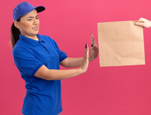 Repartidor joven en uniforme azul y gorra que se niega a tomar un paquete de papel de pie sobre una pared rosa