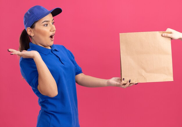 Repartidor joven en uniforme azul y gorra sintiéndose emocionada mientras recibe el paquete de papel de pie sobre la pared rosa