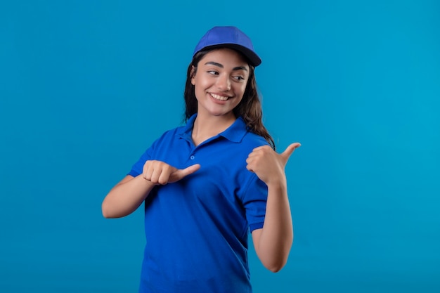 Repartidor joven en uniforme azul y gorra sonriendo confiado apuntando hacia el lado con los pulgares de pie sobre fondo azul.