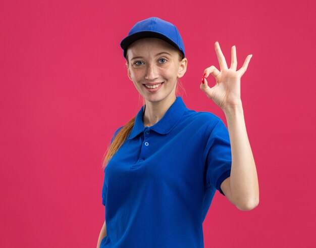 Repartidor joven en uniforme azul y gorra sonriendo confiado haciendo bien firmar parado sobre pared rosa