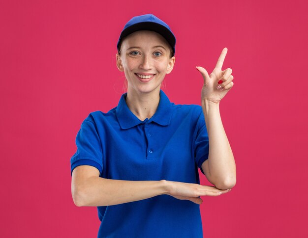 Repartidor joven en uniforme azul y gorra sonriendo confiado mostrando el dedo índice de pie sobre la pared rosa