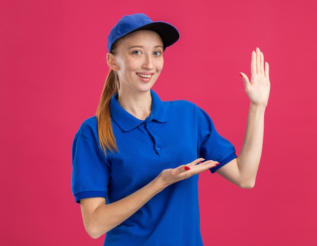 Repartidor joven en uniforme azul y gorra sonriendo confiado presentando espacio de copia con el brazo de la mano de pie sobre la pared rosa