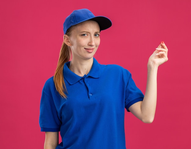 Repartidor joven en uniforme azul y gorra sonriendo haciendo gesto de dinero frotando los dedos de pie sobre la pared rosa