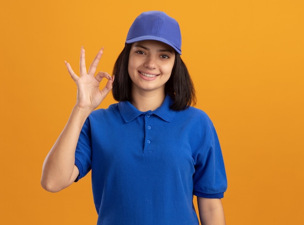 Repartidor joven en uniforme azul y gorra sonriendo mostrando ok signo de pie sobre la pared naranja