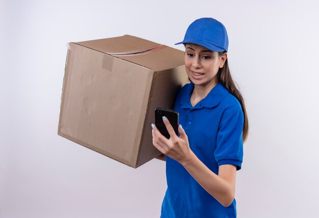 Repartidor joven en uniforme azul y gorra sosteniendo una caja de cartón grande mirando la pantalla del teléfono móvil preocupado y confundido