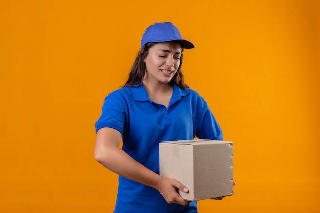 Repartidor joven en uniforme azul y gorra sosteniendo una caja de cartón mirando neumático y aburrido de pie sobre fondo amarillo