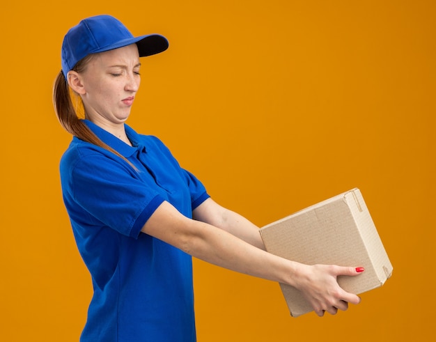 Repartidor joven en uniforme azul y gorra sosteniendo una caja de cartón mirándola con expresión de disgusto de pie sobre la pared naranja