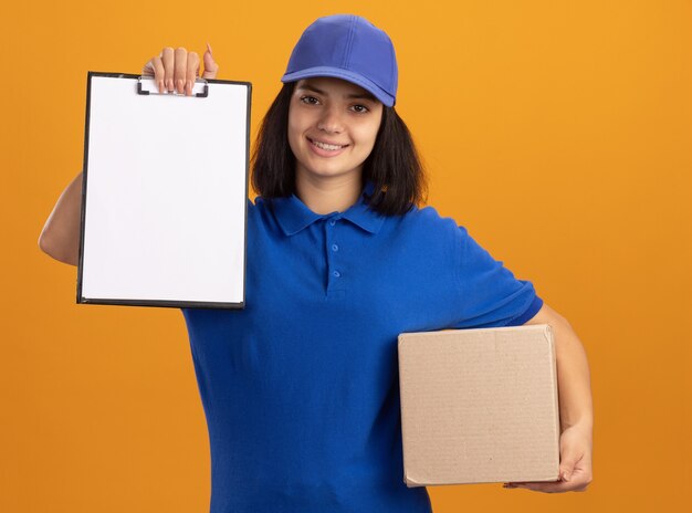 Repartidor joven en uniforme azul y gorra sosteniendo una caja de cartón que muestra el portapapeles con páginas en blanco sonriendo confiado de pie sobre la pared naranja