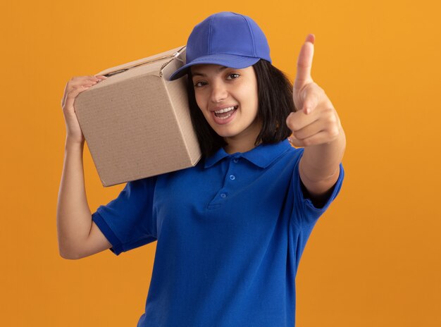 Repartidor joven en uniforme azul y gorra sosteniendo una caja de cartón sonriendo alegremente apuntando con el dedo índice de pie sobre la pared naranja