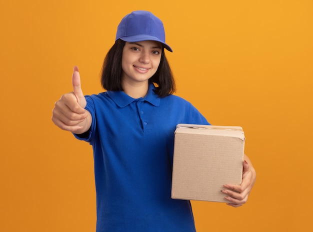 Repartidor joven en uniforme azul y gorra sosteniendo una caja de cartón sonriendo alegremente mostrando los pulgares para arriba de pie sobre la pared naranja