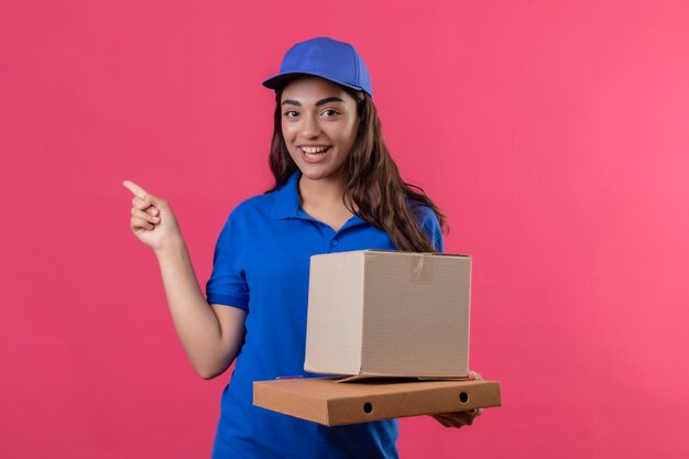 Repartidor joven en uniforme azul y gorra sosteniendo cajas de cartón apuntando con el dedo hacia el lado sonriendo alegremente feliz y positivo de pie sobre fondo rosa