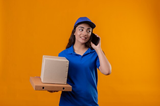 Repartidor joven en uniforme azul y gorra sosteniendo cajas de cartón hablando por teléfono móvil mirando confiado de pie sobre fondo amarillo