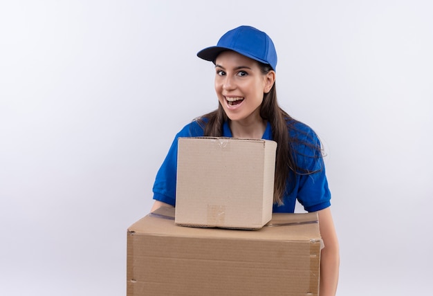 Foto gratuita repartidor joven en uniforme azul y gorra sosteniendo cajas de cartón mirando a la cámara sonriendo ampliamente, feliz y salido