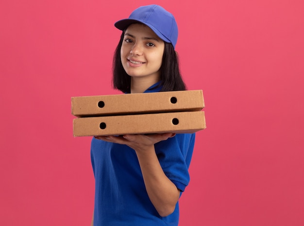 Repartidor joven en uniforme azul y gorra sosteniendo cajas de pizza con cara feliz sonriendo de pie sobre la pared rosa