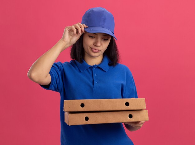 Repartidor joven en uniforme azul y gorra sosteniendo cajas de pizza haciendo boca torcida con expresión decepcionada de pie sobre la pared rosa