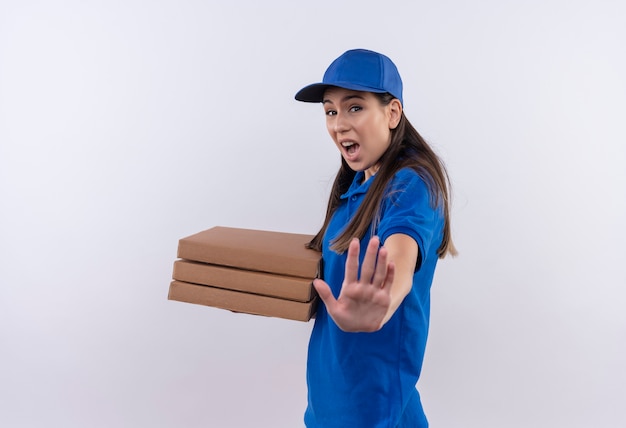 Repartidor joven en uniforme azul y gorra sosteniendo cajas de pizza haciendo señal de pare con la mano con expresión de miedo en la cara