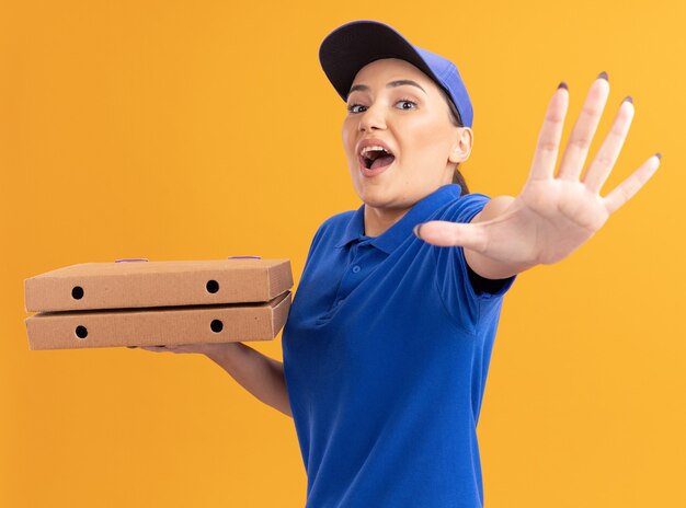 Repartidor joven en uniforme azul y gorra sosteniendo cajas de pizza mirando al frente haciendo gesto de parada con la mano preocupada de pie sobre la pared naranja