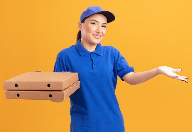 Repartidor joven en uniforme azul y gorra sosteniendo cajas de pizza mirando al frente sonriendo con cara feliz presentando con el brazo de su mano de pie sobre la pared naranja