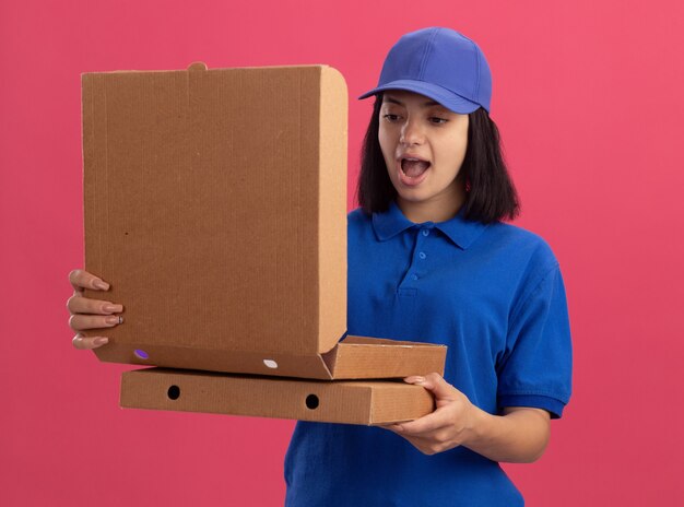 Repartidor joven en uniforme azul y gorra sosteniendo cajas de pizza mirando una caja abierta de pie asombrado y sorprendido sobre la pared rosa
