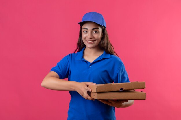 Repartidor joven en uniforme azul y gorra sosteniendo cajas de pizza mirando a la cámara sonriendo confiada feliz y positiva de pie sobre fondo rosa