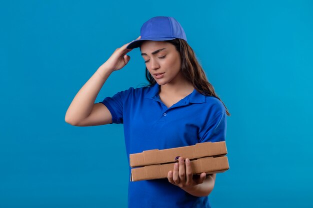 Repartidor joven en uniforme azul y gorra sosteniendo cajas de pizza mirando mal tocar la cabeza sufriendo de dolor de cabeza de pie sobre fondo azul